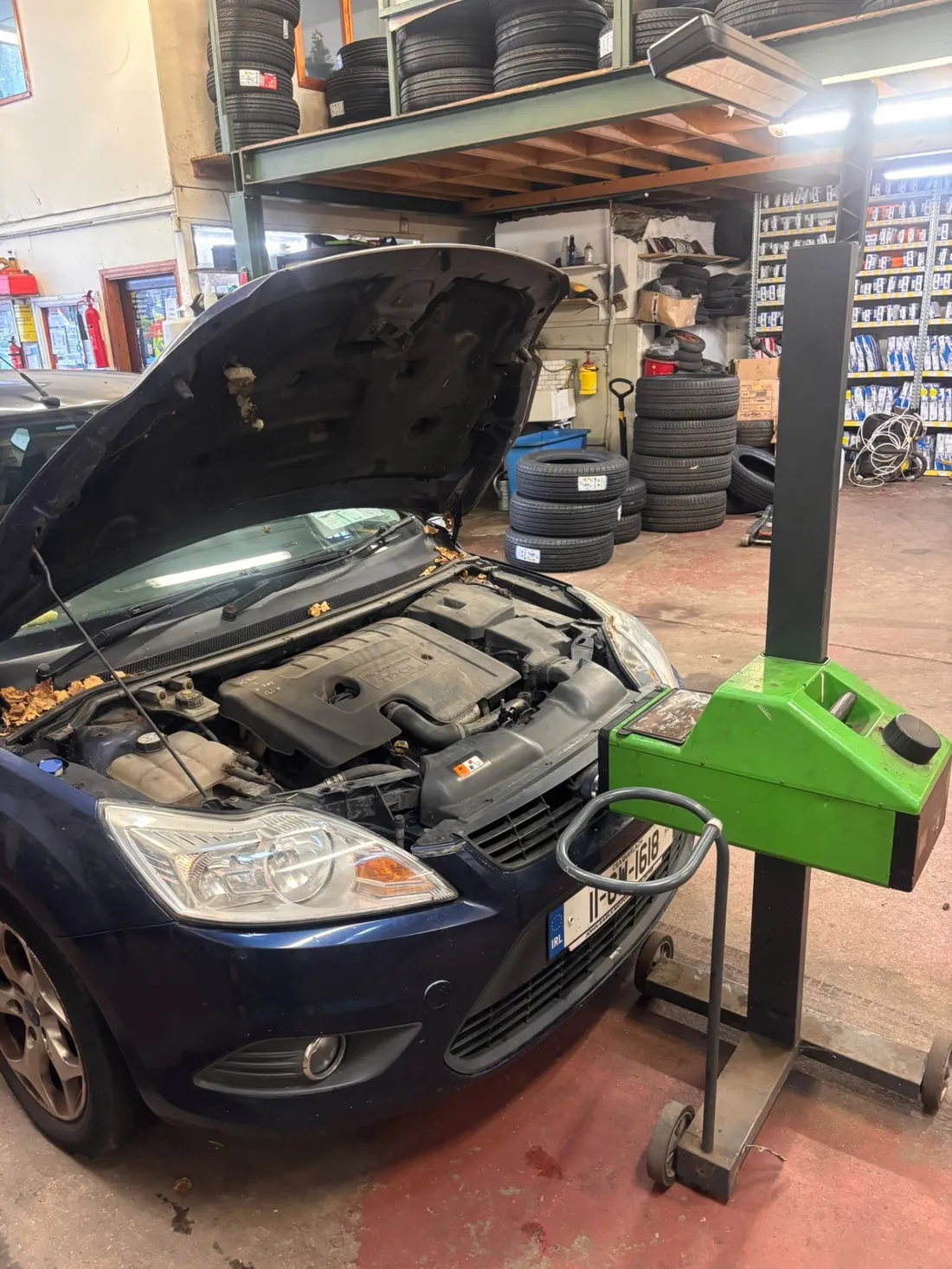 Car with bonnet open beside headlamp testing equipment, with tyres stocked on shelves behind